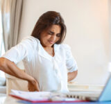Portrait of young stressed woman sitting at home office desk in front of laptop, touching aching back with pained expression, suffering from backache after working on laptop