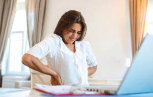 Portrait of young stressed woman sitting at home office desk in front of laptop, touching aching back with pained expression, suffering from backache after working on laptop