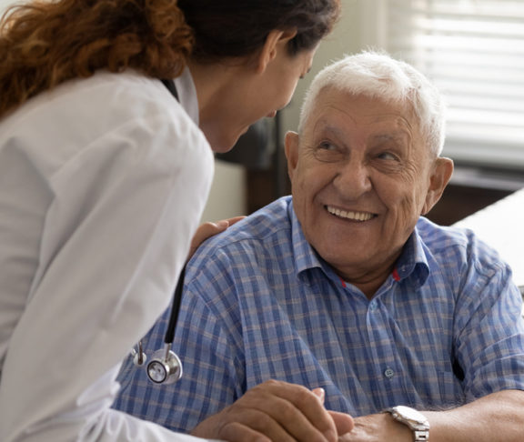 Friendly young female doctor and optimistic elder 80s man meeting in office for consultation. GP therapist holding hands of old patient, giving sympathy, support, advice about treatment