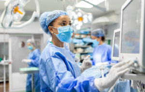 Anesthetist Working In Operating Theatre Wearing Protecive Gear checking monitors while sedating patient before surgical procedure in hospital