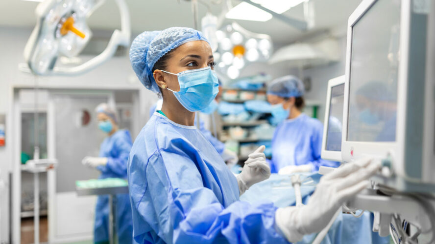 Anesthetist Working In Operating Theatre Wearing Protecive Gear checking monitors while sedating patient before surgical procedure in hospital