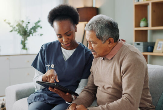 Nurse, senior and discussion with tablet on sofa for healthcare, communication or consultation at home. Woman or medical caregiver helping and talking to elderly male person on technology for advice