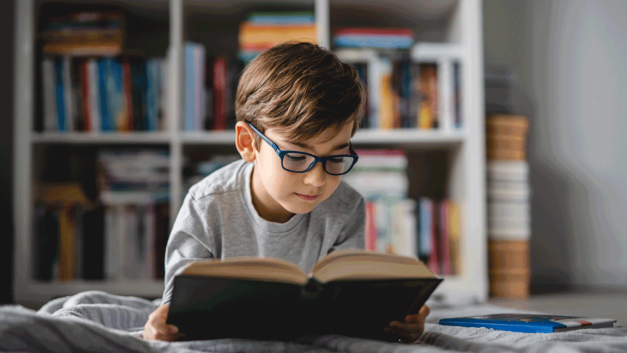 One caucasian boy lying on the floor at home in day reading a book front view wearing eyeglasses copy space real people