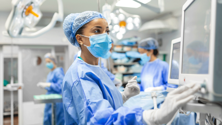 Anesthetist Working In Operating Theatre Wearing Protecive Gear checking monitors while sedating patient before surgical procedure in hospital